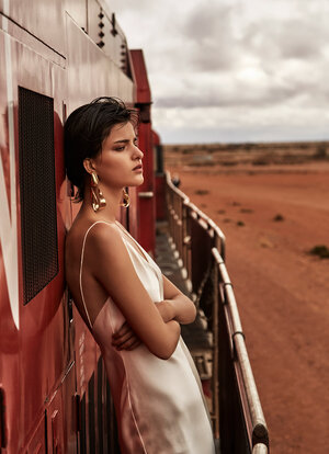 A brunette woman in a white dress stands on a train platform with her arms crossed, gazing toward a dusty desert landscape under cloudy skies.