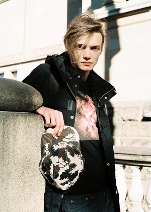 A fair-skinned young man with blond hair in a black jacket and t-shirt, holding a printed baseball cap, leaning against a stone wall.
