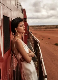 Woman in a white slip dress leaning against a red train car in the desert, featuring hair styling by Kyye