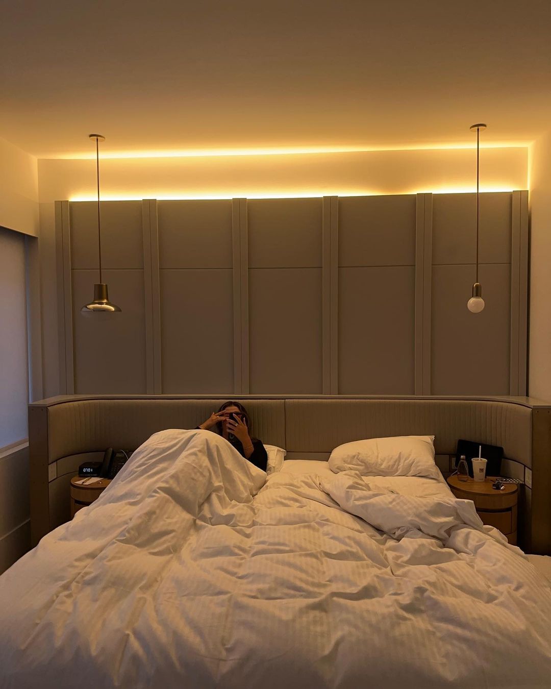Person in bed with white duvet and pillows, bedside tables, and wall lighting in the neutral-toned bedroom interior.