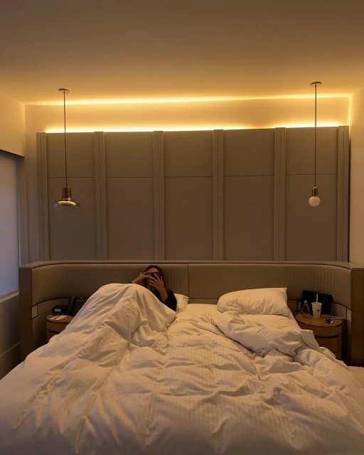 Person in bed with white duvet and pillows, bedside tables, and wall lighting in the neutral-toned bedroom interior.