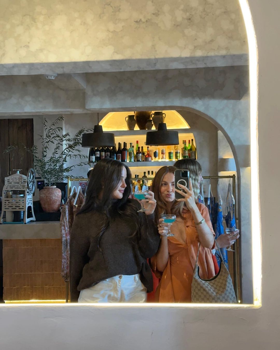 Three young adult women pose in front of a mirror at a bar with cocktails, capturing a selfie with a smartphone.