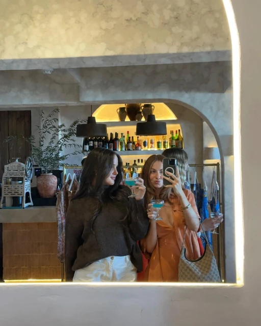 Three young adult women pose in front of a mirror at a bar with cocktails, capturing a selfie with a smartphone.