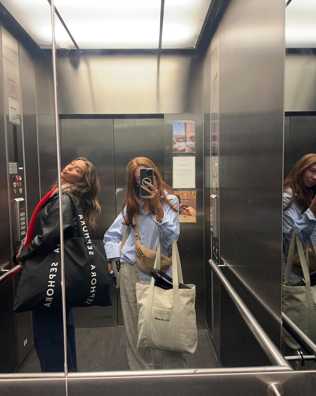 Two young women pose for a selfie in an elevator with stainless steel walls, holding shopping bags, under bright overhead lighting.