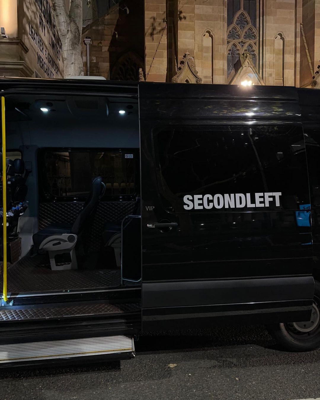 Black van with its sliding door open, revealing a glimpse of the interior, parked in an urban setting at night, illuminated by street lights.