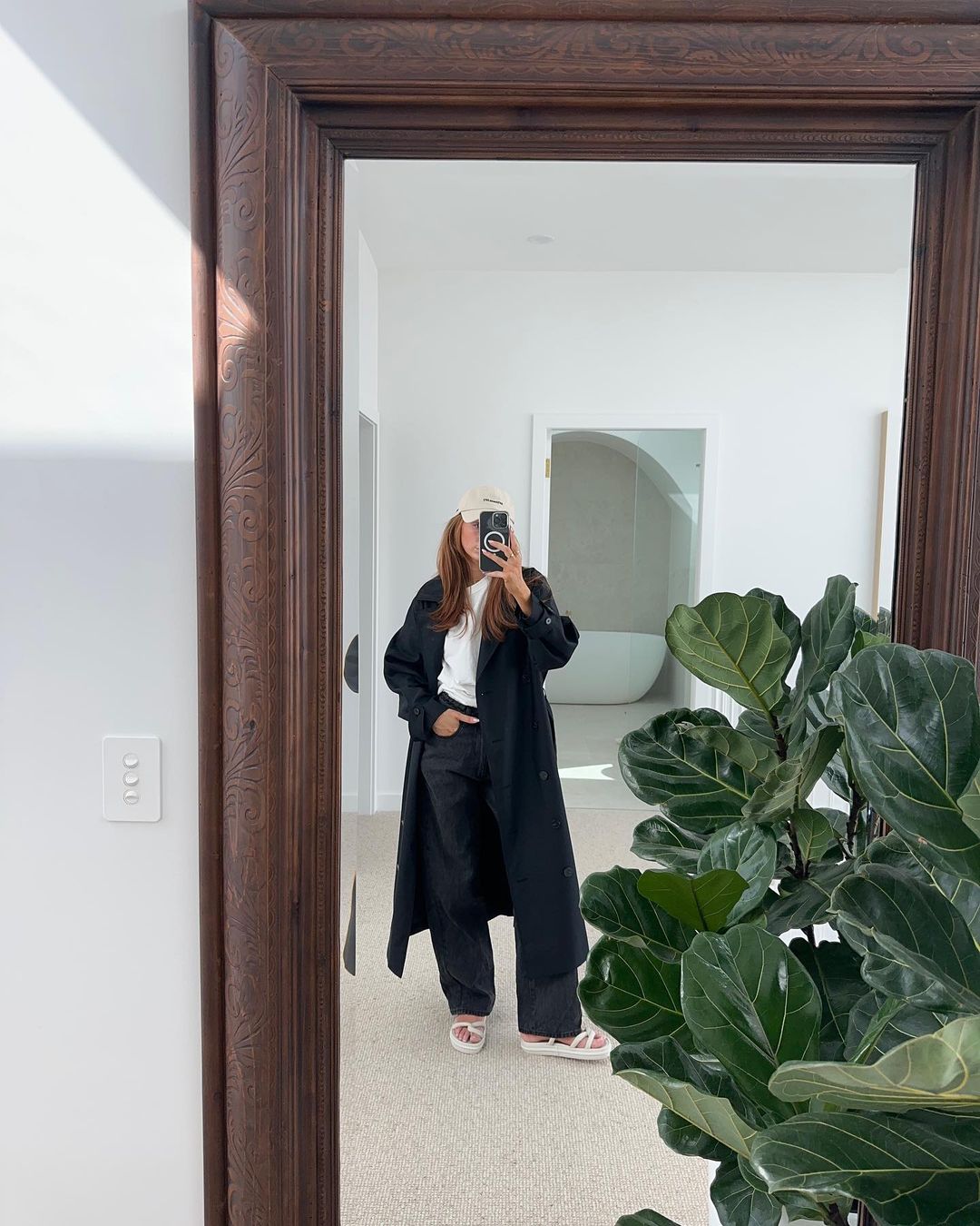 A woman takes a mirror selfie indoors, dressed in a long black coat and light-colored sandals, with a potted plant nearby.