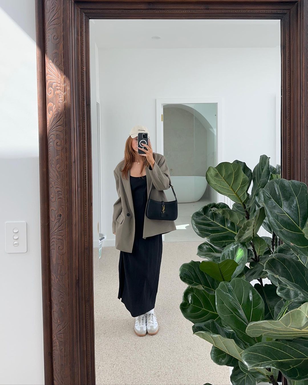 Woman in tan blazer, black dress and white sneakers takes mirror selfie in a minimalist white interior, showcasing modern fashion.