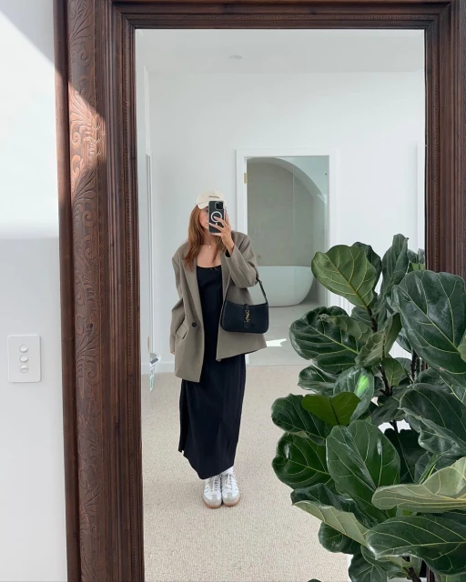 Woman in tan blazer, black dress and white sneakers takes mirror selfie in a minimalist white interior, showcasing modern fashion.