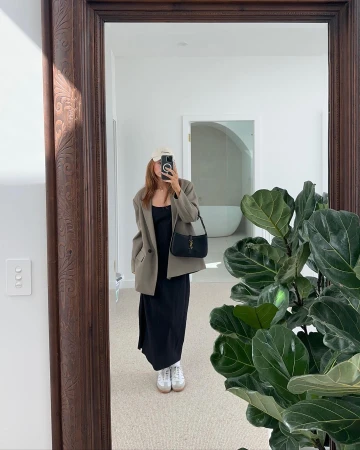 Woman in tan blazer, black dress and white sneakers takes mirror selfie in a minimalist white interior, showcasing modern fashion.