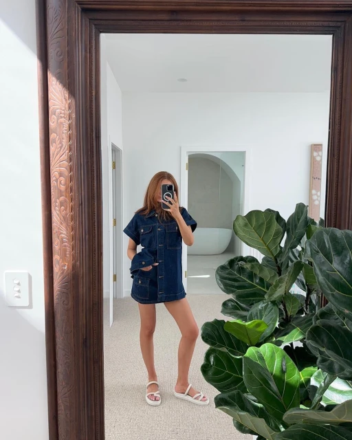 Adult female poses in denim dress, reflected in mirror with ornamental wood frame, with potted plant, bathroom and white walls.