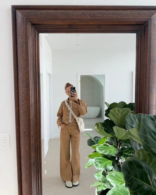 A woman in a brown denim jacket and pants takes a mirror selfie indoors, with a plant and a glimpse of a white bathroom.