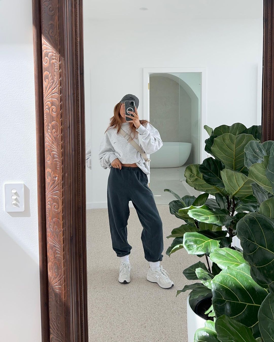 Woman taking a mirror selfie wearing an oversized grey sweatshirt and dark sweatpants in a bright room with a large potted plant