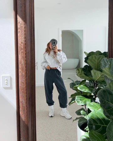 Woman taking a mirror selfie wearing an oversized grey sweatshirt and dark sweatpants in a bright room with a large potted plant