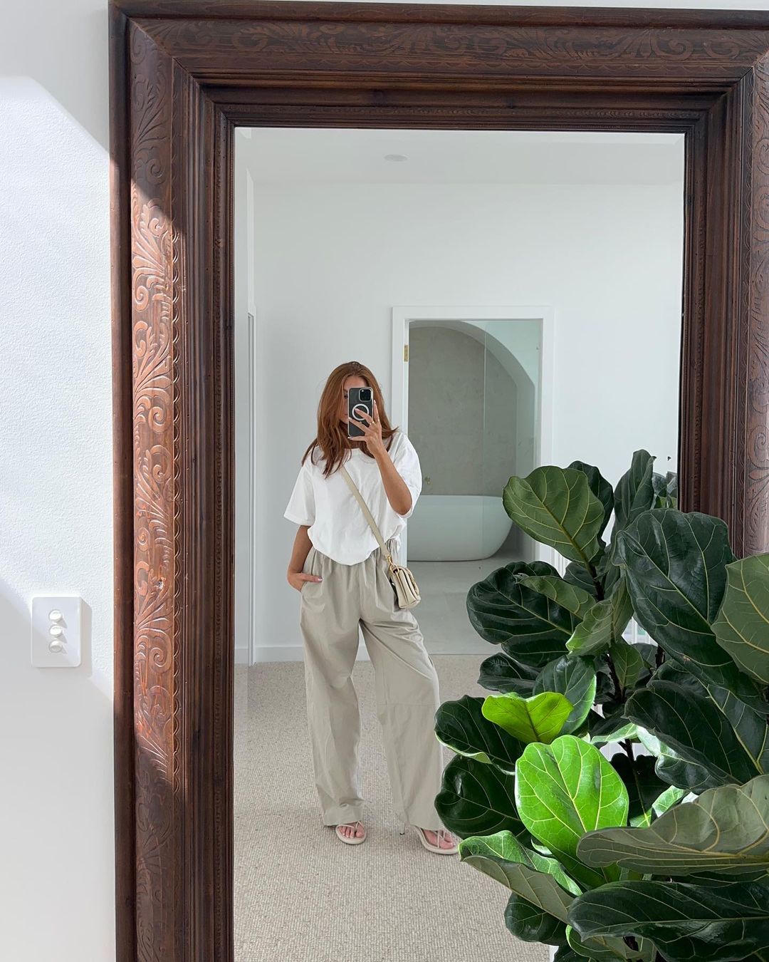 Woman in mirror selfie with oversized white t-shirt and beige pants, shoulder bag, indoor with plant, arch doorway.