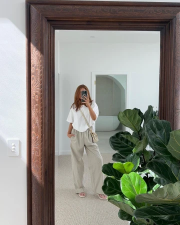 Woman in mirror selfie with oversized white t-shirt and beige pants, shoulder bag, indoor with plant, arch doorway.