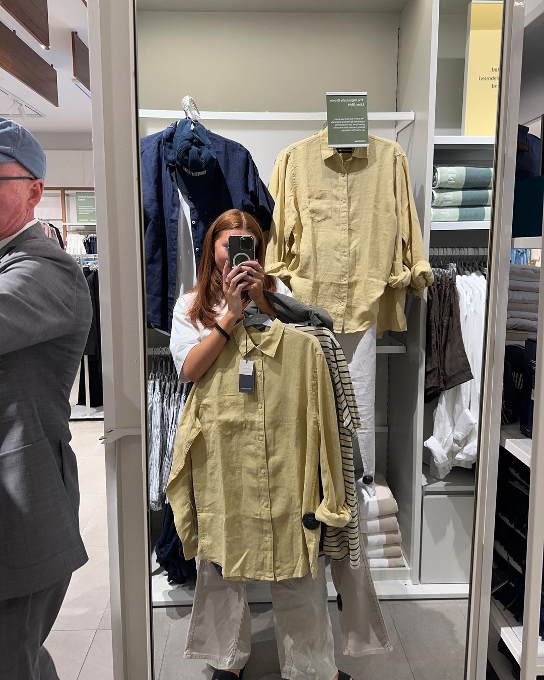 A woman takes a mirror selfie holding a yellow button-down shirt and wearing wide-leg pants in a clothing store.