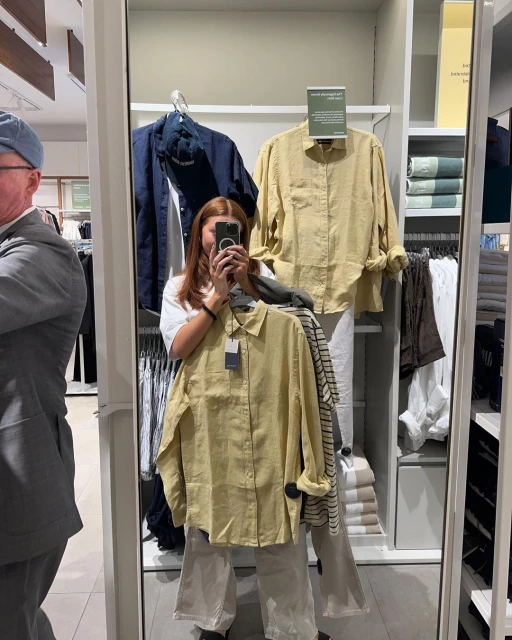 A woman takes a mirror selfie holding a yellow button-down shirt and wearing wide-leg pants in a clothing store.
