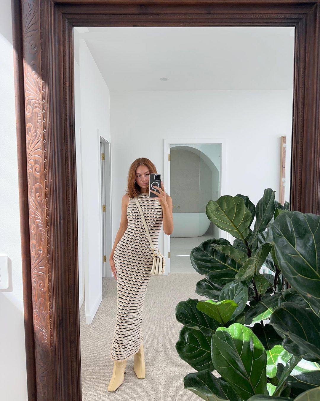 Woman in striped dress takes a mirror selfie inside a white-walled home interior, featuring a large green plant and framed mirror.