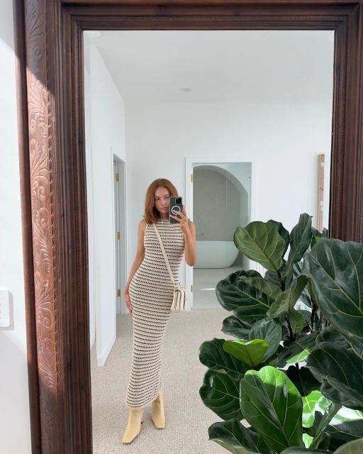 Woman in striped dress takes a mirror selfie inside a white-walled home interior, featuring a large green plant and framed mirror.