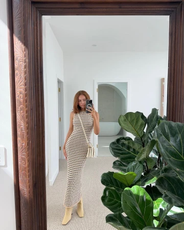 Woman in striped dress takes a mirror selfie inside a white-walled home interior, featuring a large green plant and framed mirror.