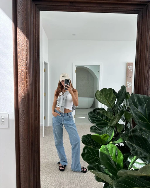 Woman takes a mirror selfie in casual outfit with light blue jeans, a white tank top, and a tan baseball cap in a white room.