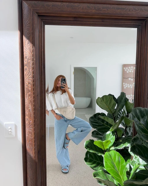 A woman poses in front of a mirror, wearing light blue jeans, a white top, and a cream-colored bag. A green plant is beside her.