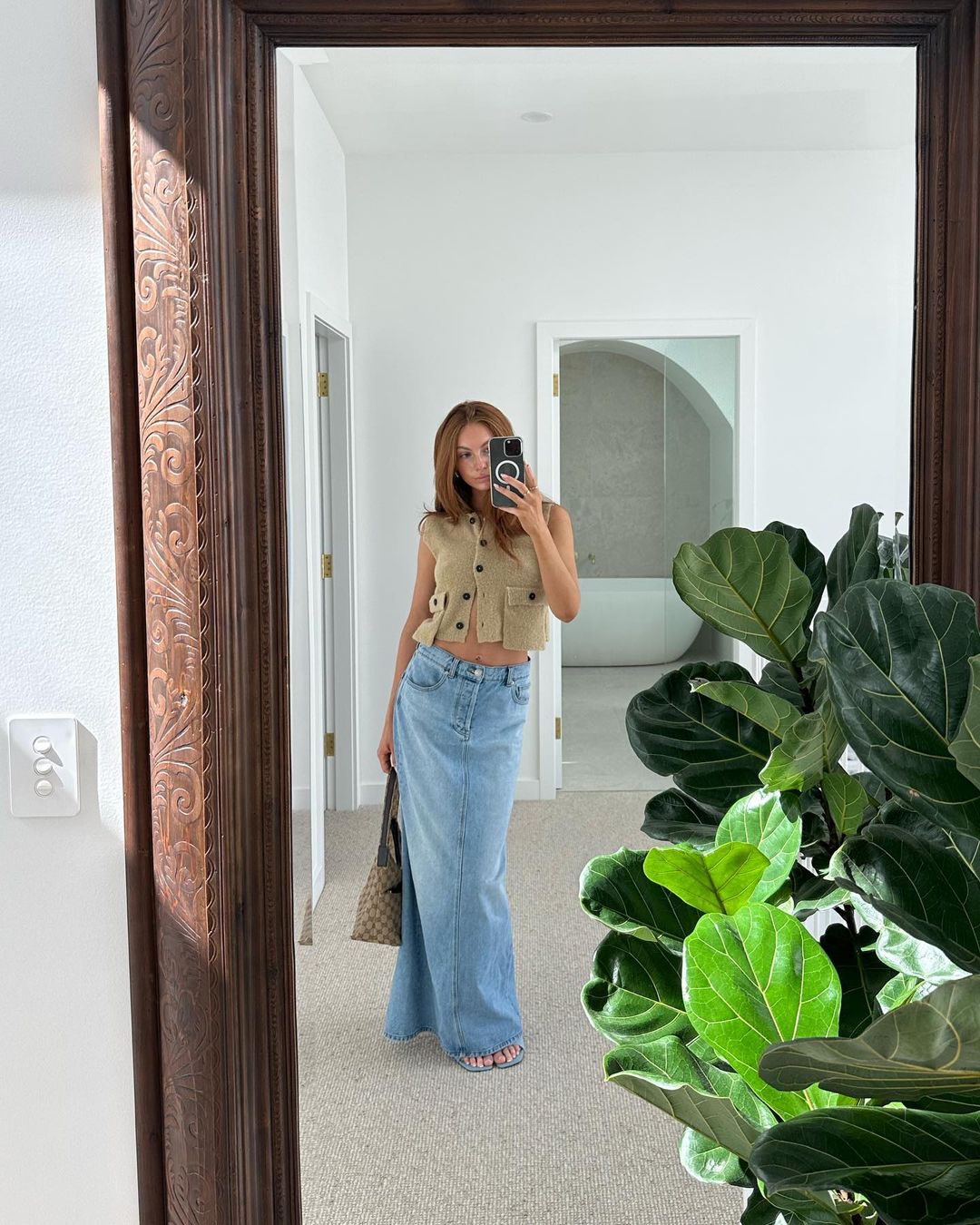 A woman takes a mirror selfie indoors, wearing a cropped beige top and long denim skirt, with a potted plant and hallway visible.