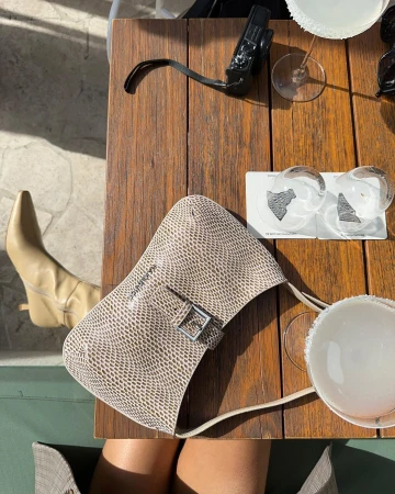 Overhead shot of a snakeskin-patterned purse on a wooden table with a tan boot, glassware, and a digital camera in soft lighting.