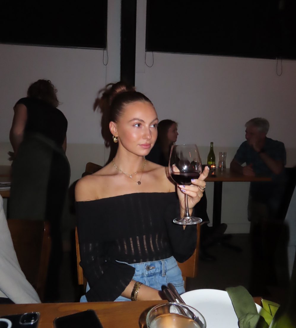 Young woman in a black off-the-shoulder top sits at a restaurant table holding a glass of red wine, with other diners in background.