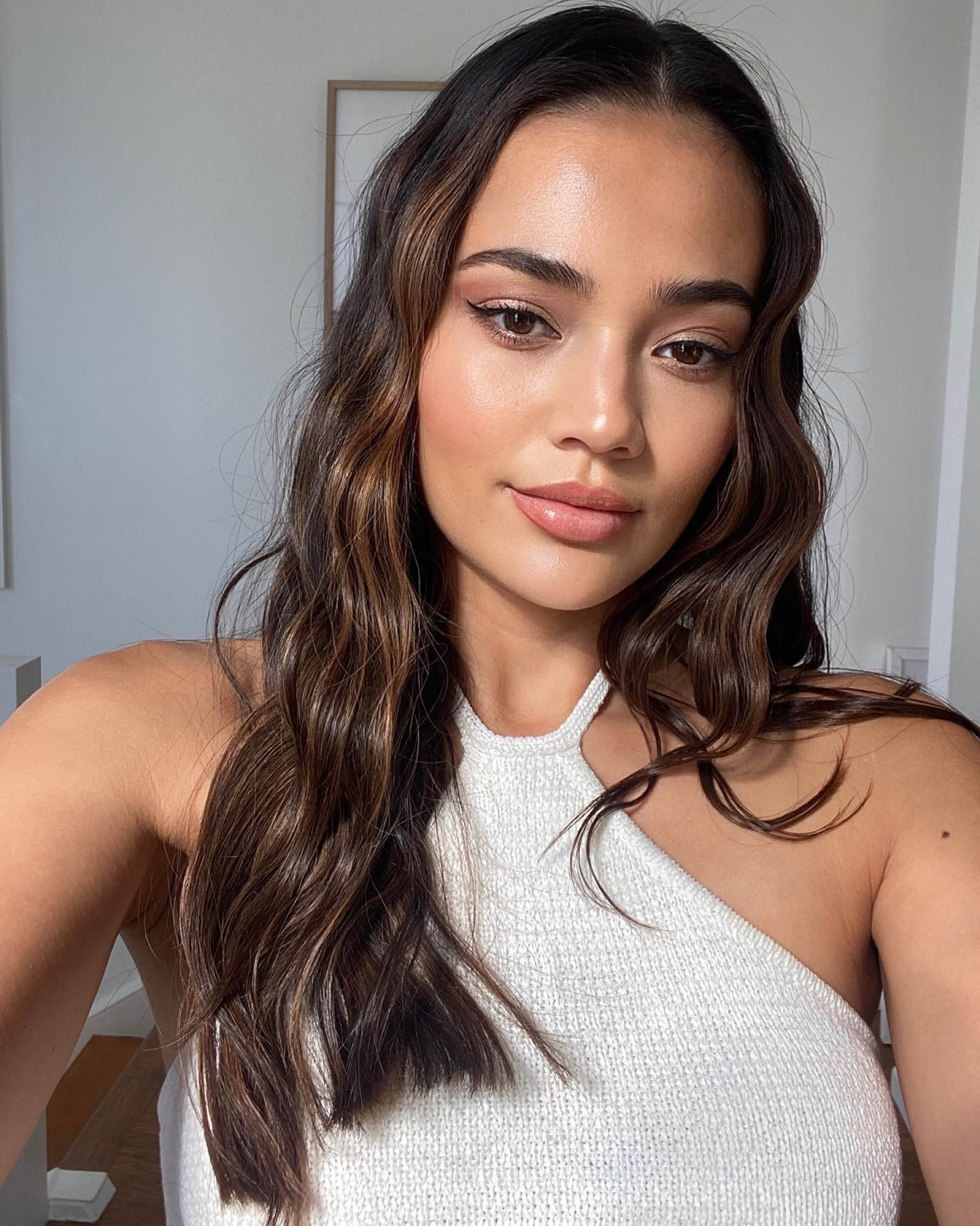 Portrait of a young woman with wavy, dark brown hair, wearing a white top, in an indoor setting with natural light.