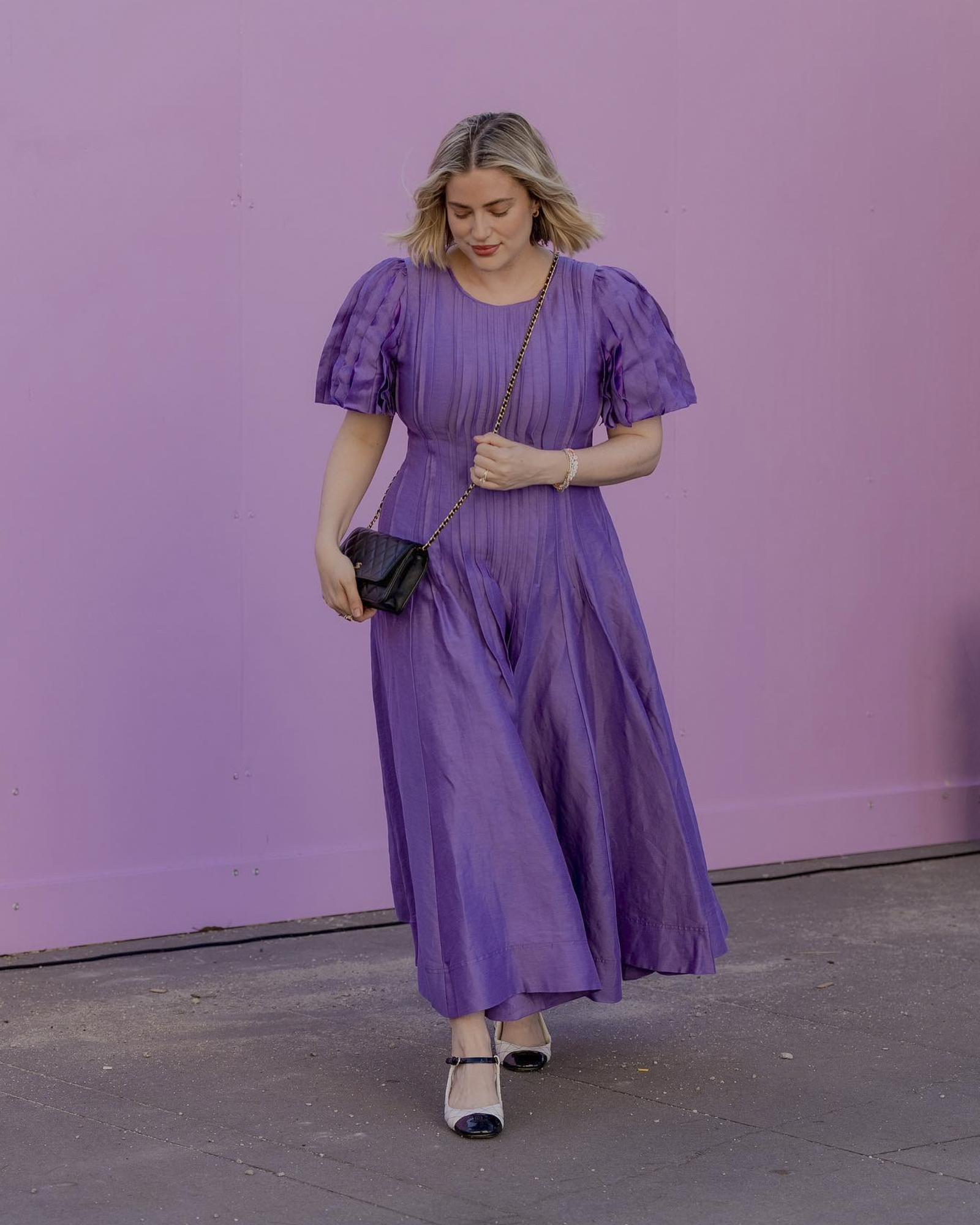 Adult female in a purple midi dress with puffed sleeves and a black purse posing against a purple wall in an urban setting.