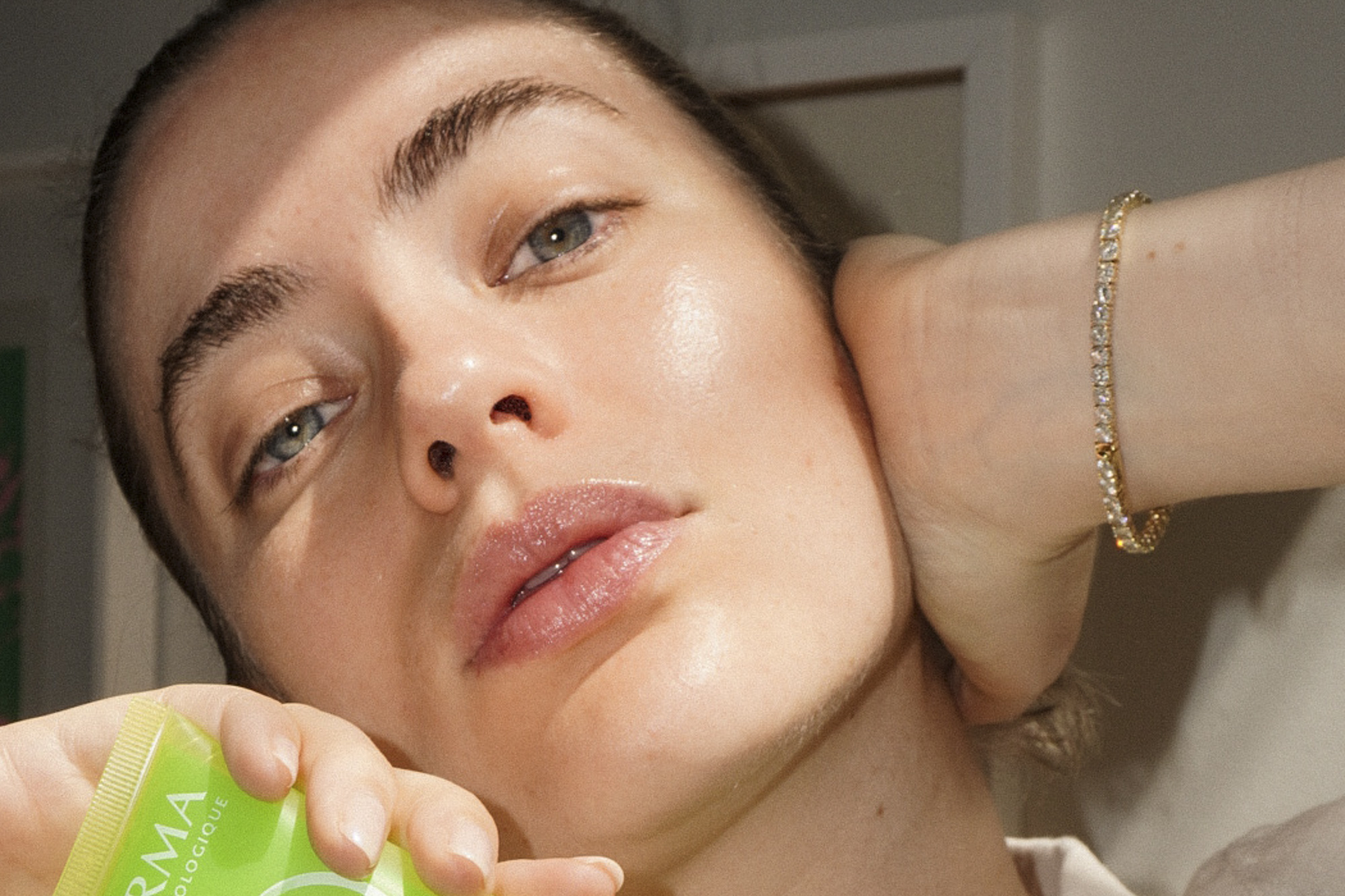 Close-up shot of a fair-skinned woman holding green skincare product, showing glossy skin, pink lips, and a diamond bracelet.