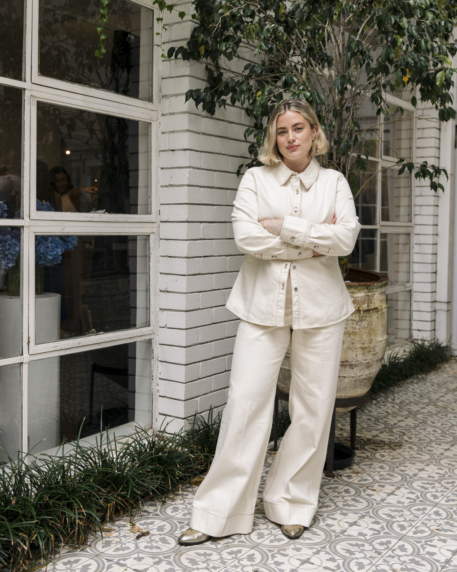 Full-length shot of a woman in white pants and a matching button-up shirt with her arms crossed in front of a white-brick building.
