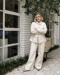 Woman in cream button-up shirt and wide-leg trousers standing outdoors with arms crossed against a white brick wall.