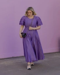 Woman wearing a pleated purple midi dress with short sleeves and holding a small black handbag against a solid pink wall