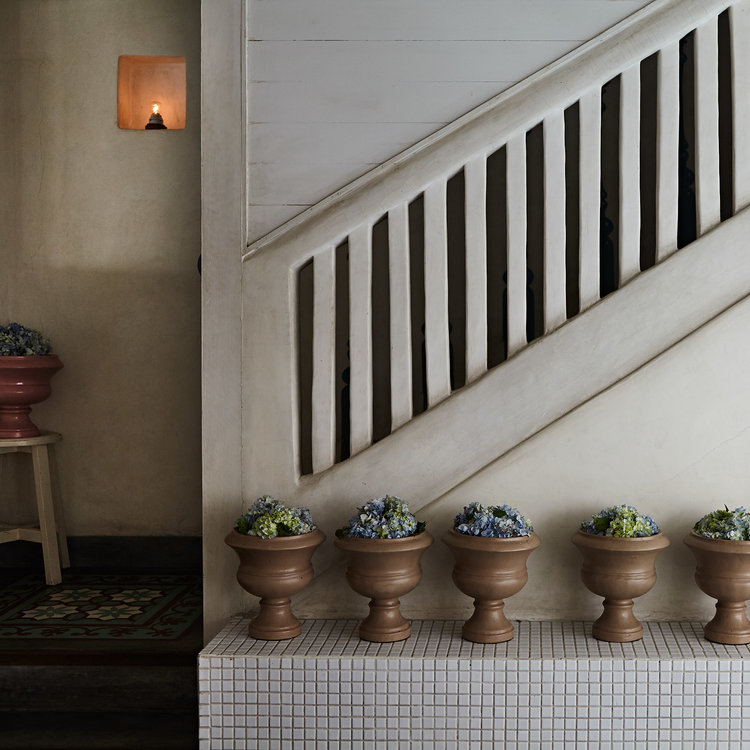 Interior view of a staircase with white banisters, accompanied by a row of terracotta pots filled with plants on a tiled surface.