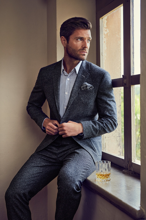 An adult man in a dark suit, shirt, and pocket square sits on a windowsill, gazing out a window with a glass of whiskey nearby.