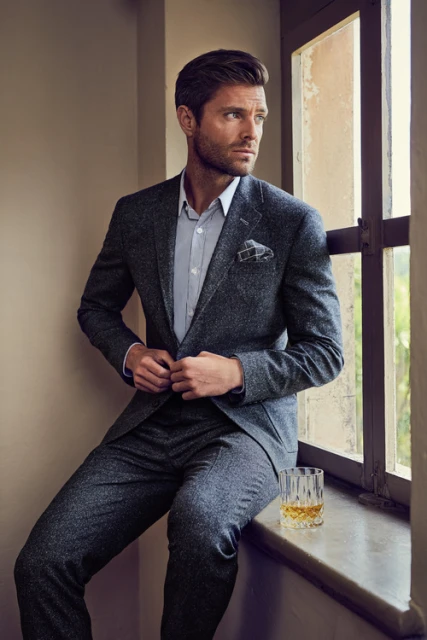An adult man in a dark suit, shirt, and pocket square sits on a windowsill, gazing out a window with a glass of whiskey nearby.