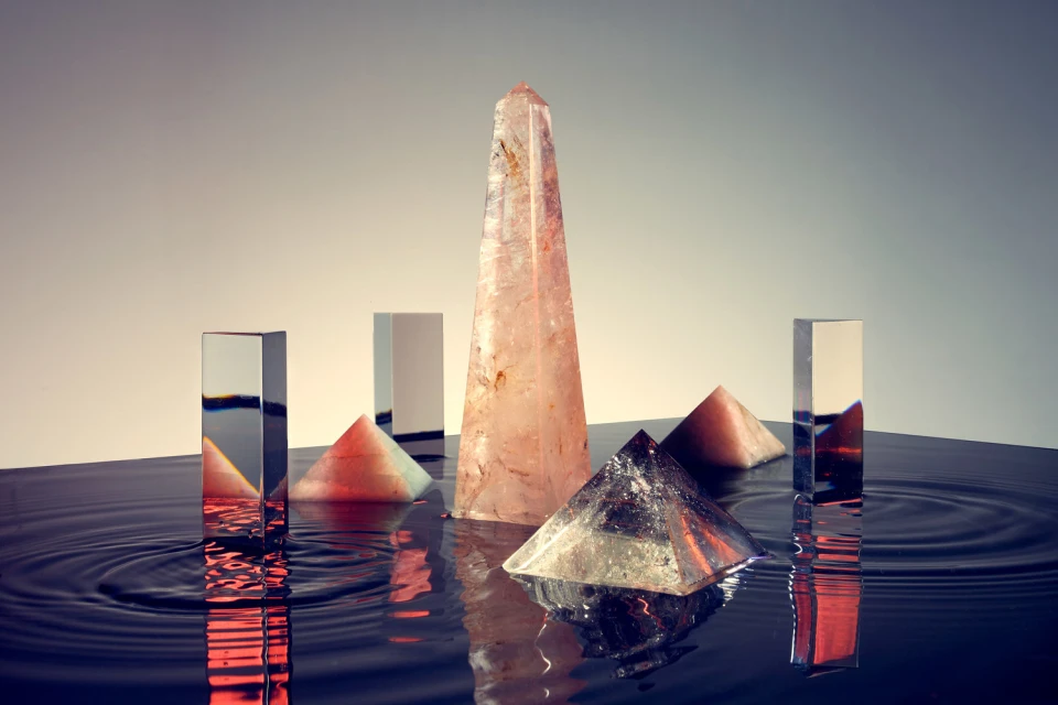 Still life featuring geometric crystal shapes including pyramids and rectangular prisms on a glossy, wet surface, shot in a photo studio.