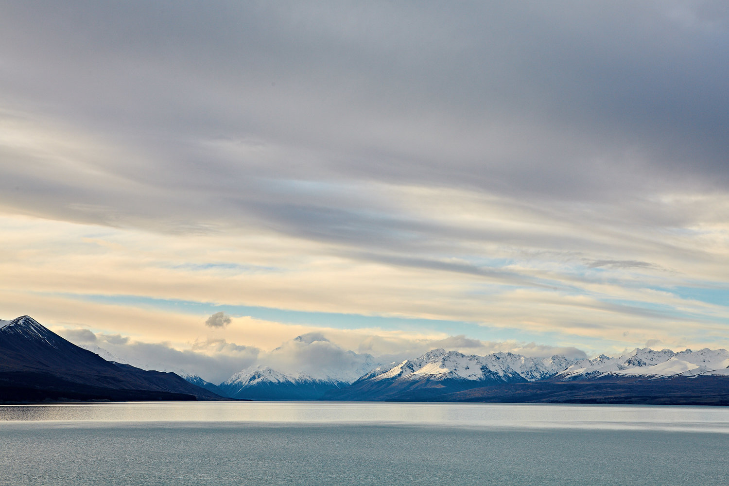 Scenic view of a calm lake reflecting snow-capped mountains under a cloudy sky, creating a peaceful and serene natural landscape.