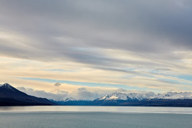 Calm body of water stretching toward snow-capped mountain peaks under an expansive cloudy sky photographed by Andrew Butler.