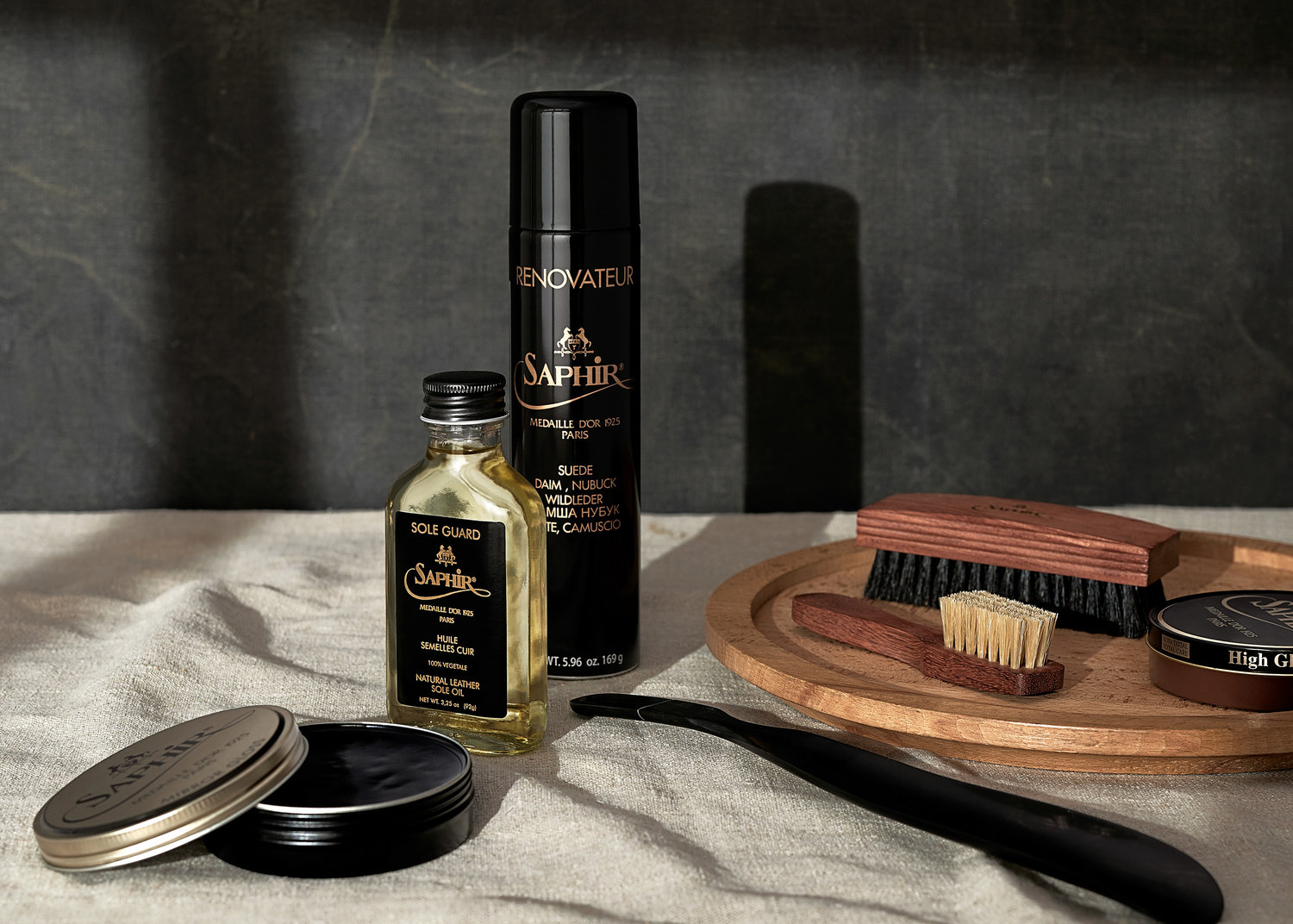 Close-up of Saphir brand shoe care products: Renovateur spray, Sole Guard oil, polish, and brushes, arranged on a neutral linen surface.