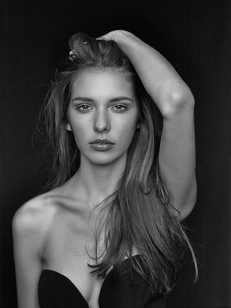 Black and white portrait of a young woman with hand in hair, wearing a strapless top against a dark backdrop in a photo studio.