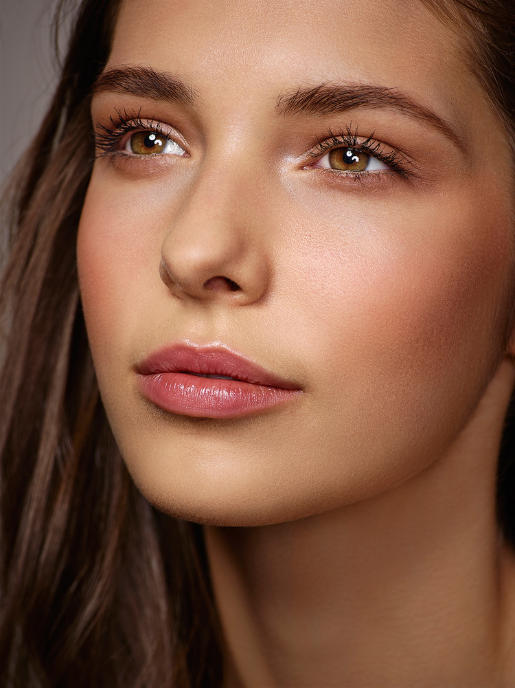 Close-up portrait of a young adult female with brown eyes and wavy brown hair, light skin, wearing pink lipstick against a gray backdrop.