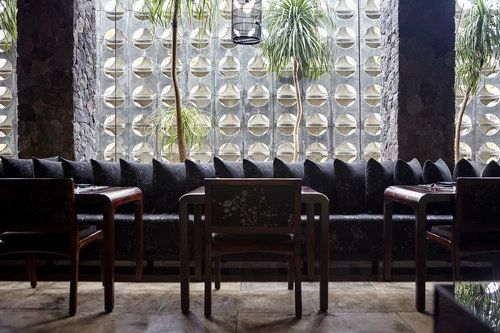 Interior shot of a restaurant featuring dark wooden tables, chairs, a long plush sofa, and a patterned concrete screen wall.