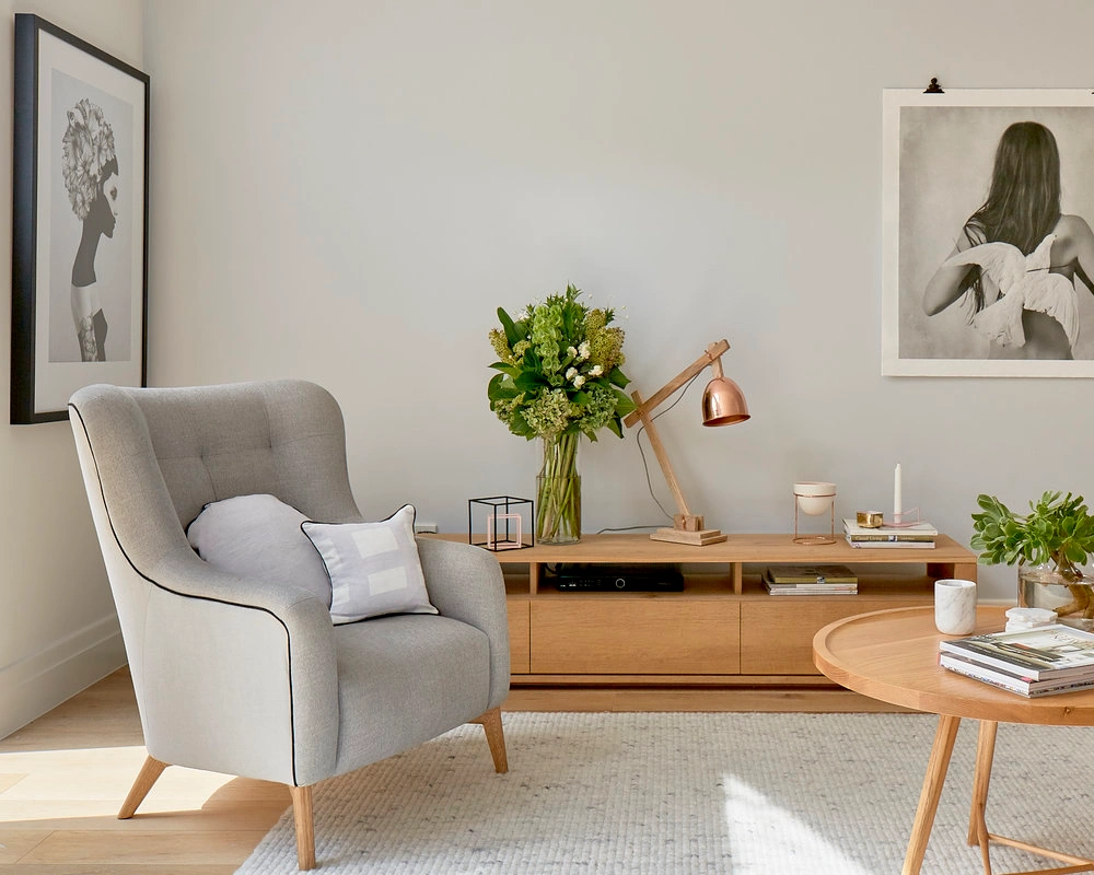 A grey upholstered armchair in a sunlit modern living room with a wooden credenza, floral arrangement, and framed wall art.