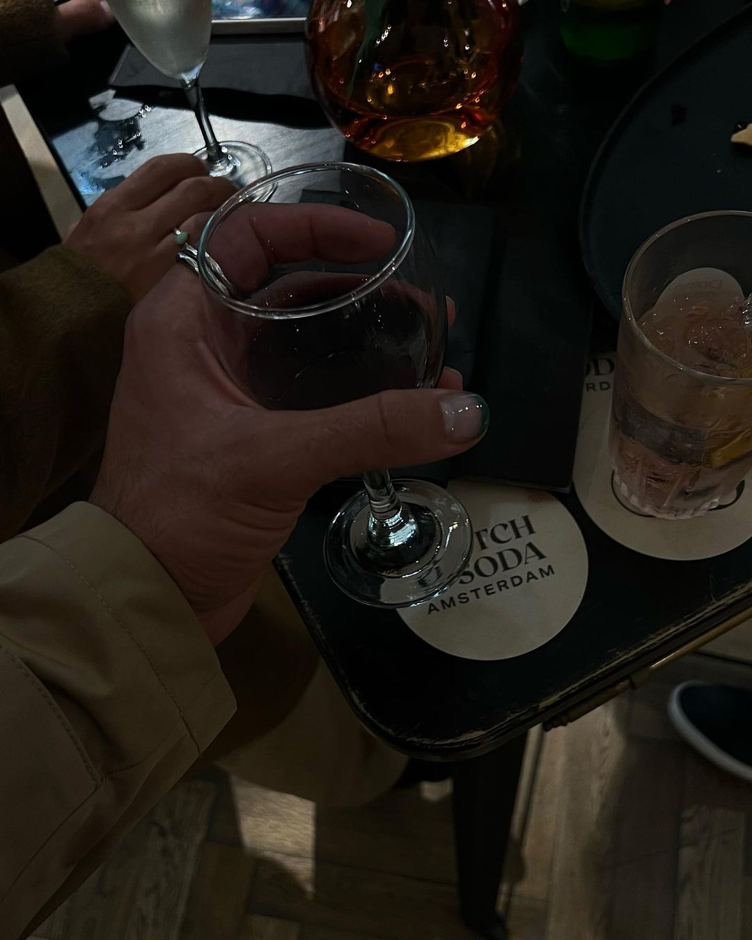 Close-up of a hand holding a glass of red wine, with another drink and hands visible in background in a dimly lit indoor setting.