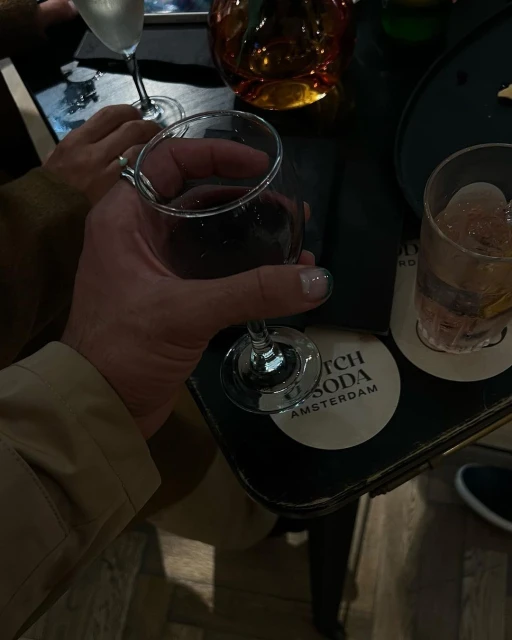 Close-up of a hand holding a glass of red wine, with another drink and hands visible in background in a dimly lit indoor setting.