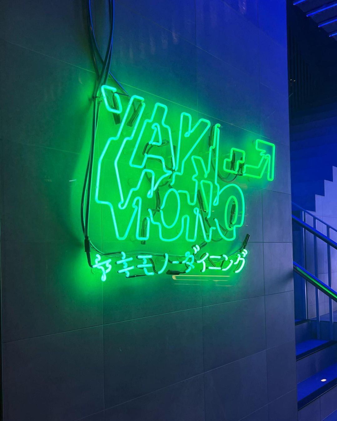 Close-up of a green neon sign that reads "Yakiniku Dining" on a tiled wall with a glimpse of stairs and blue light in the background.