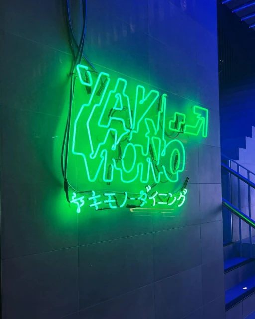 Close-up of a green neon sign that reads "Yakiniku Dining" on a tiled wall with a glimpse of stairs and blue light in the background.